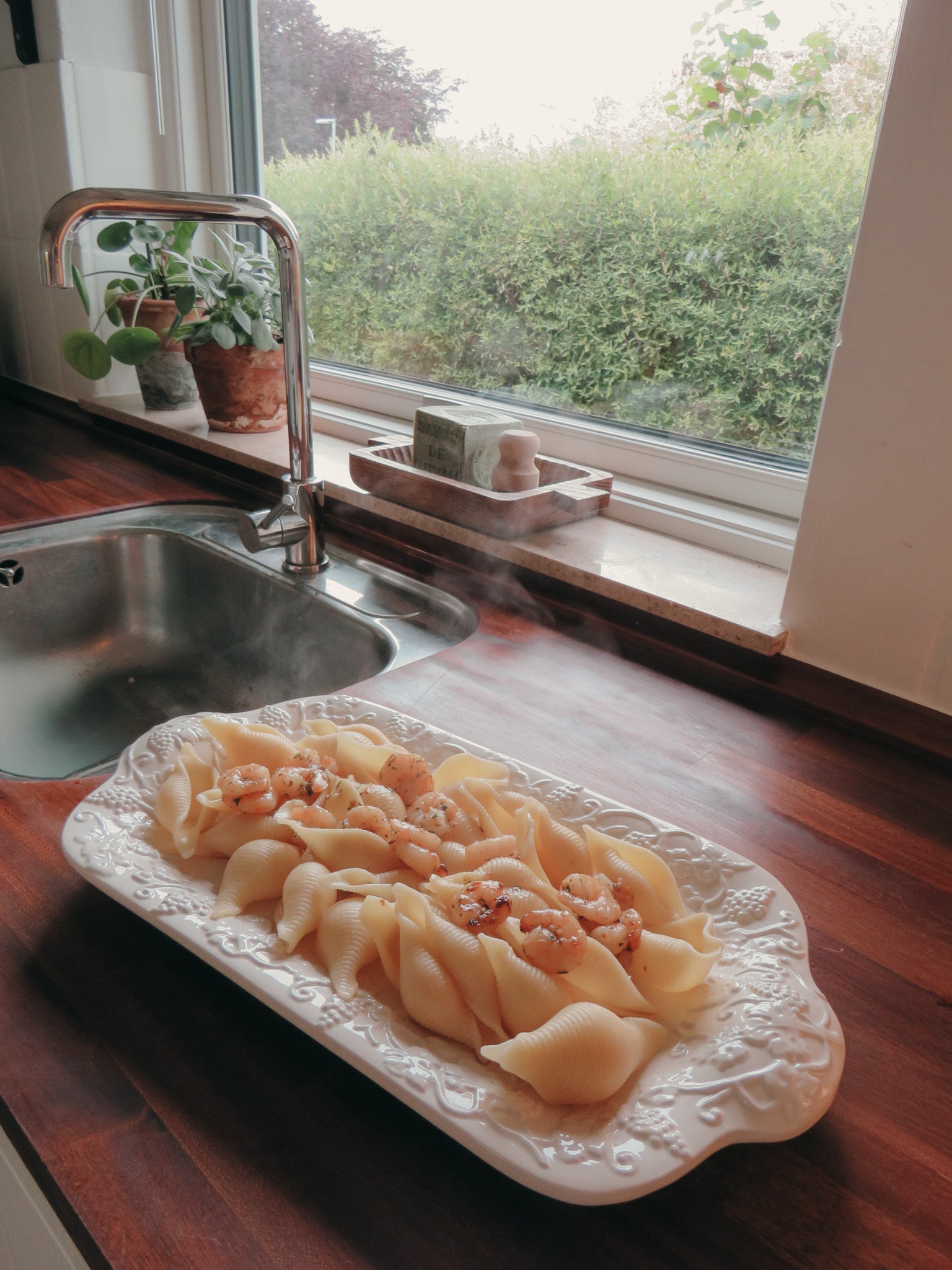 A white rectangular platter filled with pasta and topped with nuts, set on a dark wooden kitchen counter next to a sink and window.