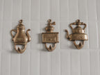 Three vintage brass wall hooks shaped like teapots and a coffee grinder, mounted on a white tiled wall.