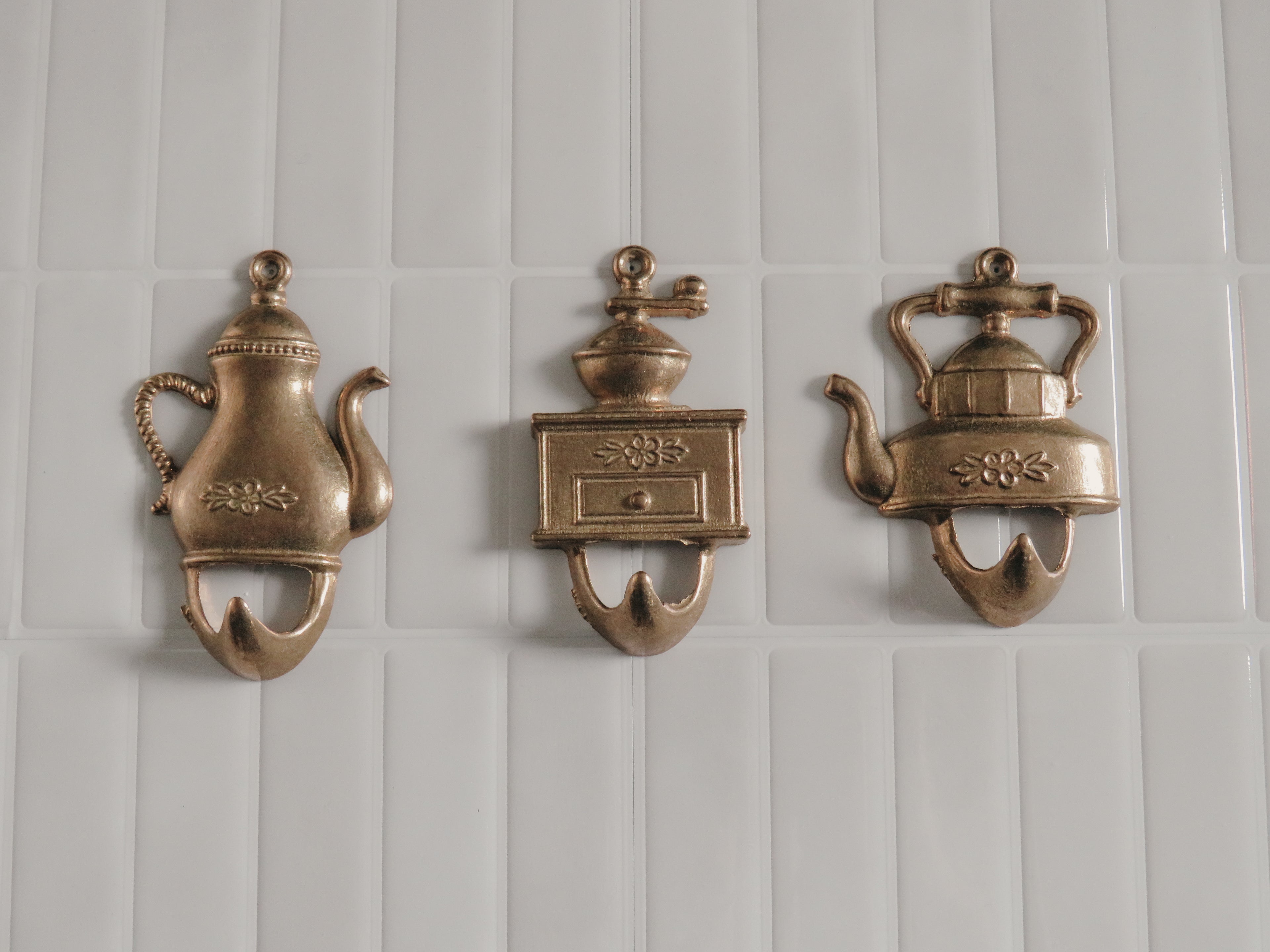 Three vintage brass wall hooks shaped like teapots and a coffee grinder, mounted on a white tiled wall.