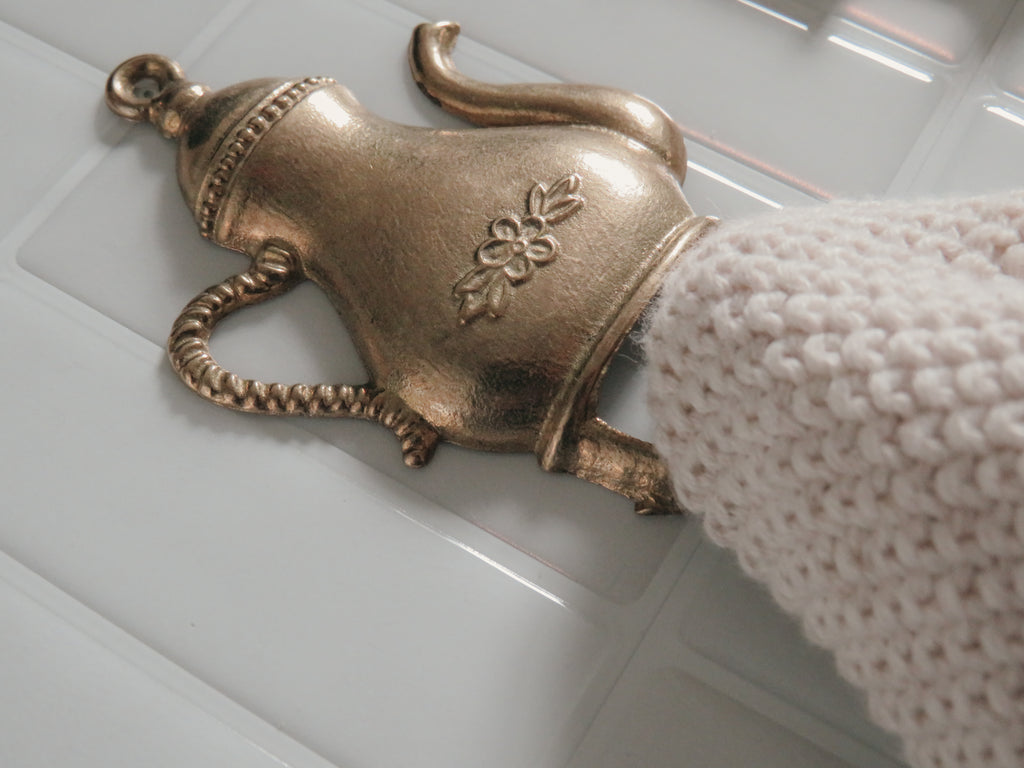 A close-up of a metallic brass teapot-shaped drawer pull handle with a floral engraving, shown installed on a white paneled door next to a white knit sleeve.
