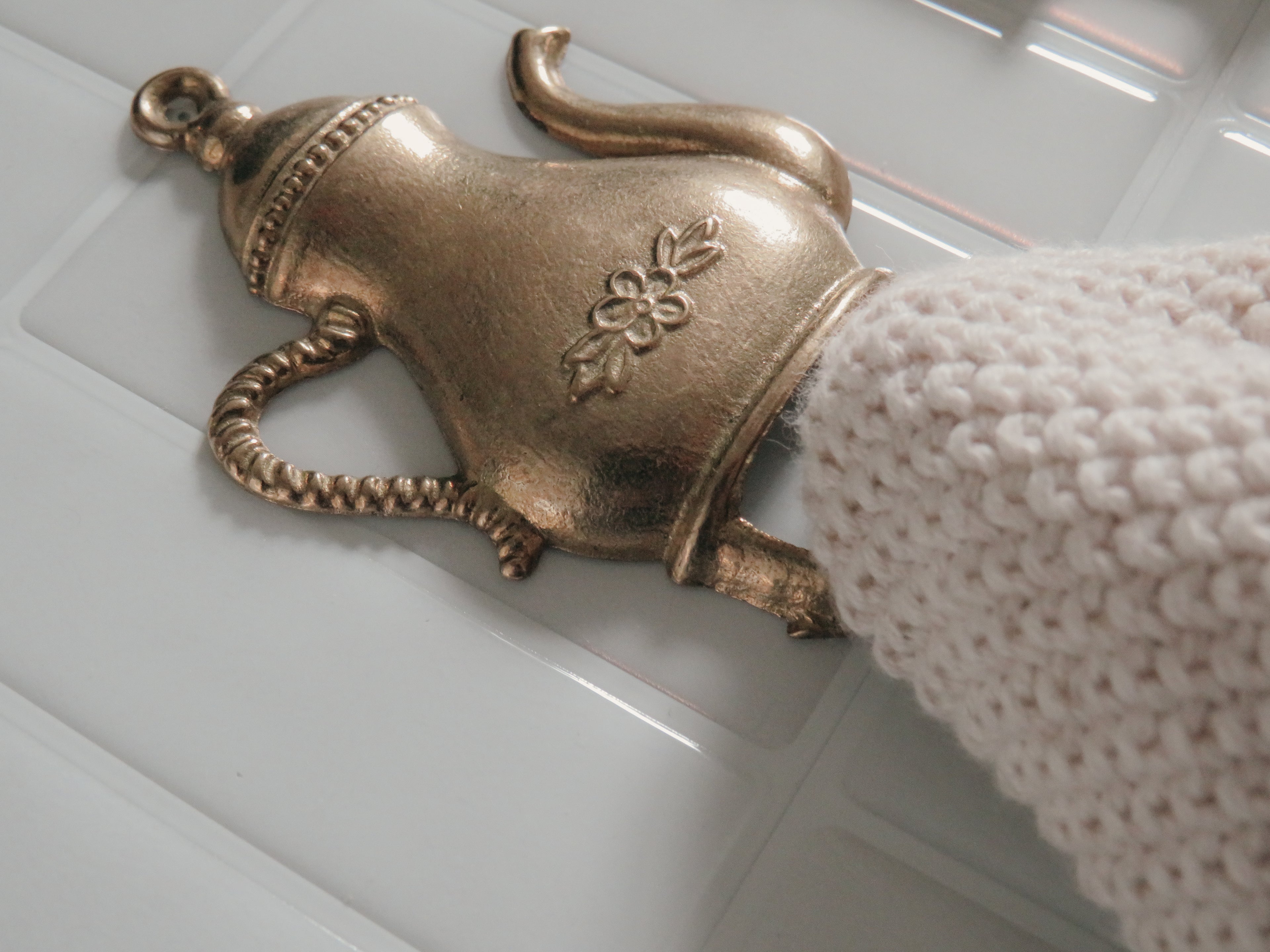 A close-up of a metallic brass teapot-shaped drawer pull handle with a floral engraving, shown installed on a white paneled door next to a white knit sleeve.