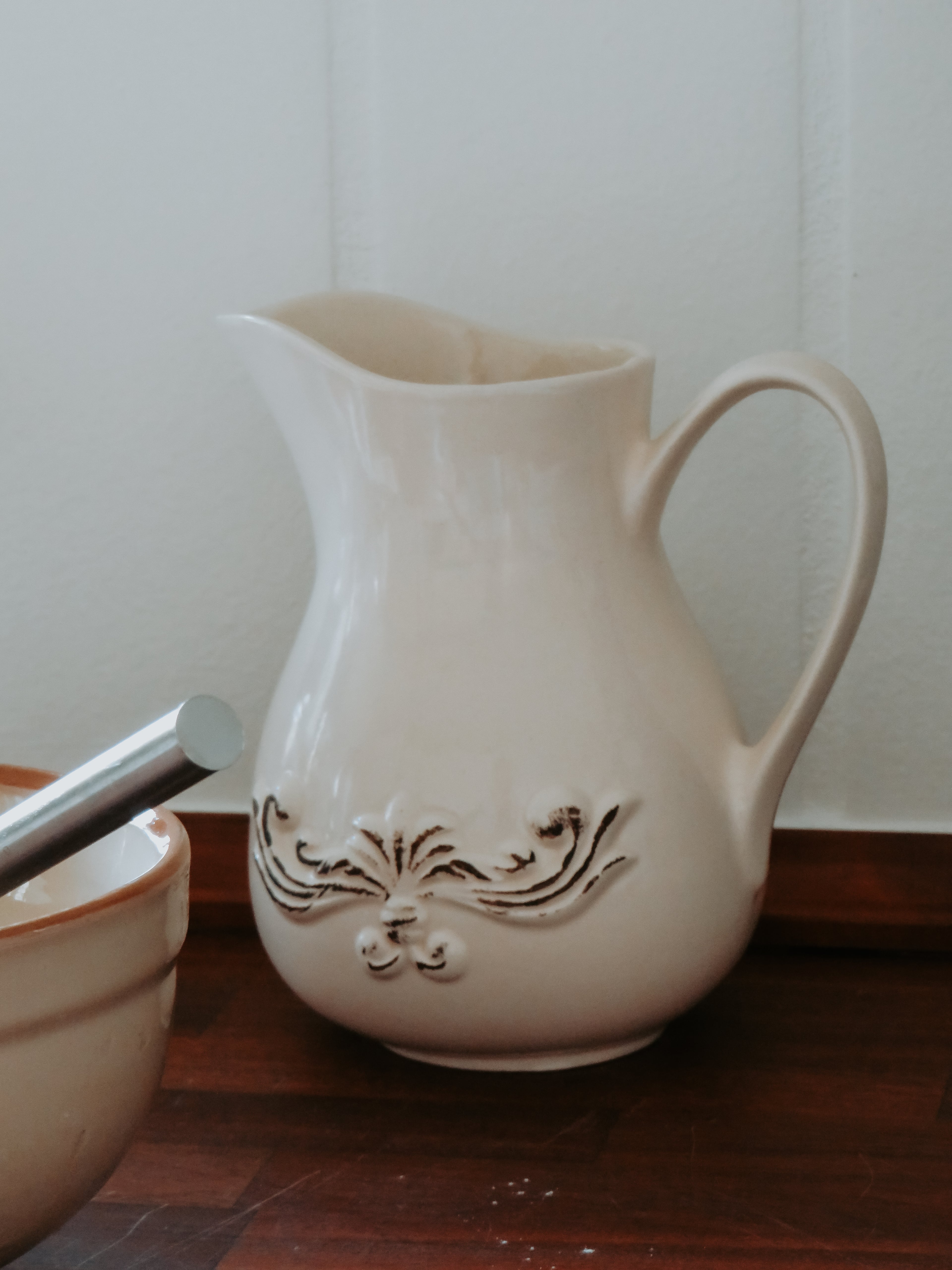 An off-white ceramic pitcher with embossed floral detailing on the front, sitting on a dark wooden surface against a white paneled wall.