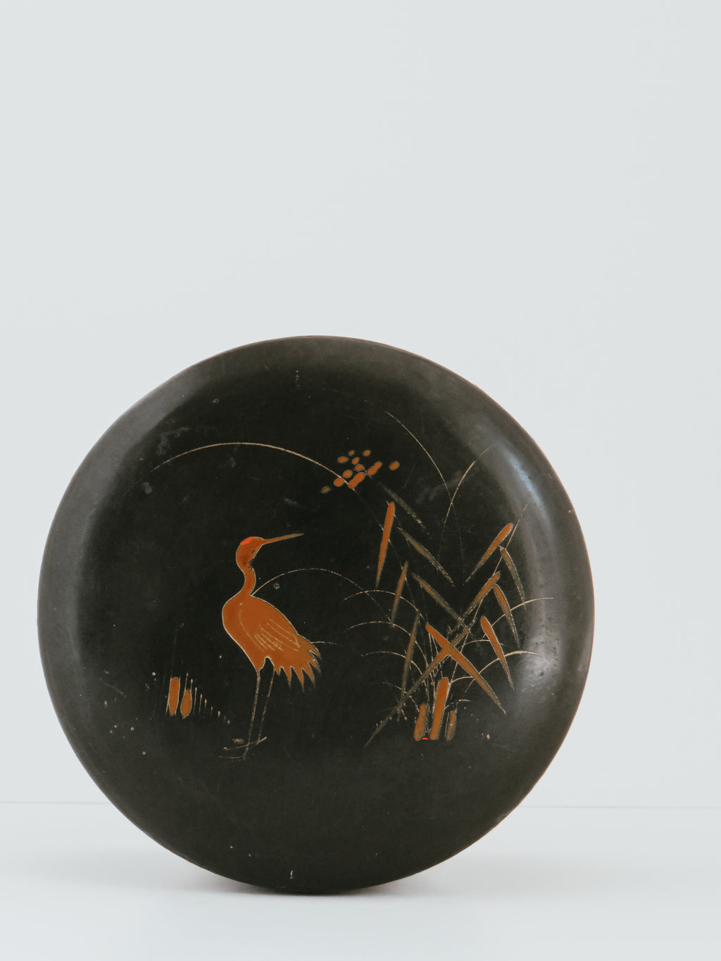 A round black lacquered box lid featuring hand-painted orange and gold illustrations of a heron and reeds, set against a plain light gray background.