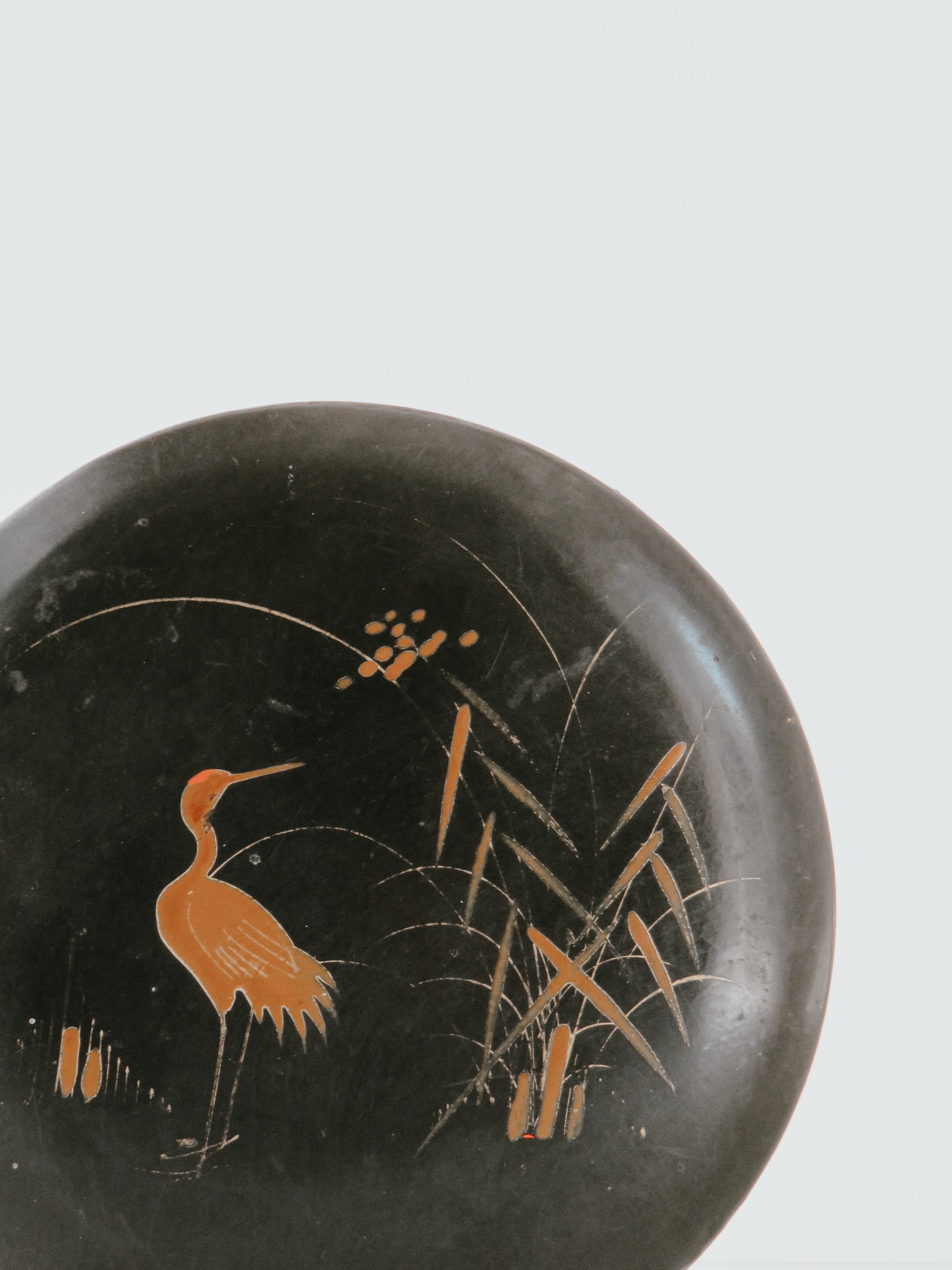 Close-up of a round black decorative object featuring a painted orange heron and reeds on a light gray background.