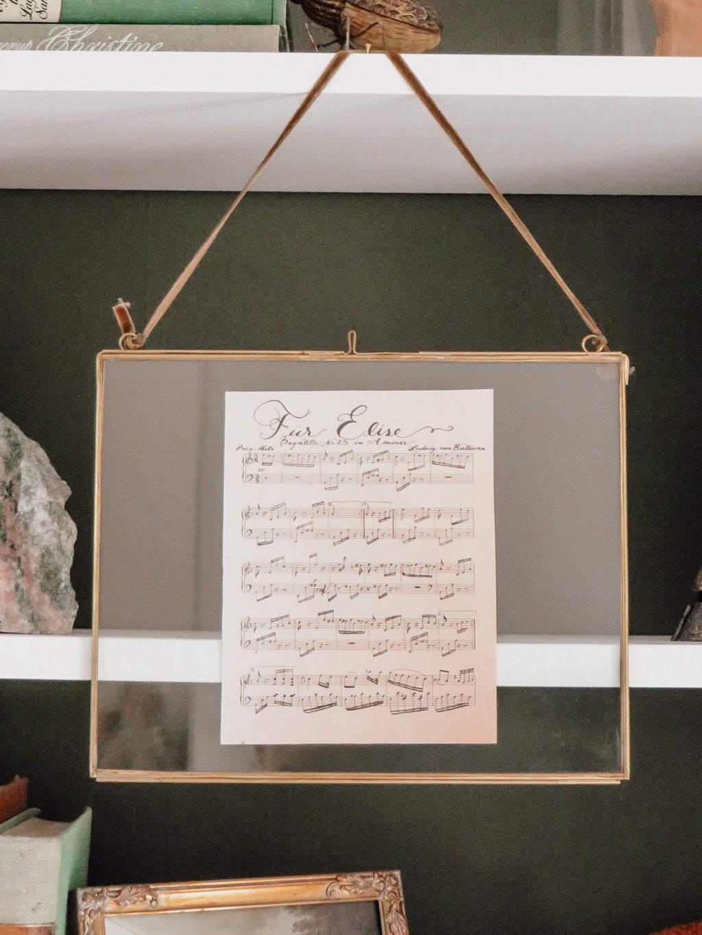 A framed hanging print with a sheet music design displayed on a shelf among books and decorative objects.