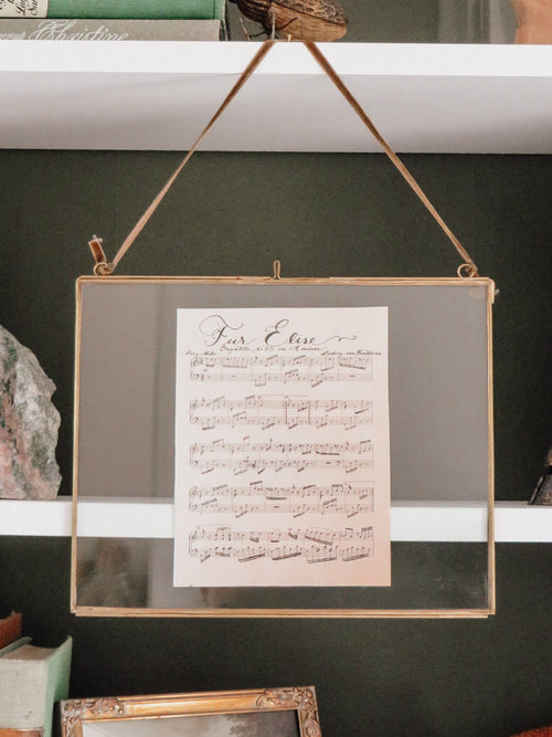 A framed hanging print with a sheet music design displayed on a shelf among books and decorative objects.