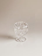 A clear cut-glass or crystal footed bowl with a lid, photographed on a warm beige studio background.