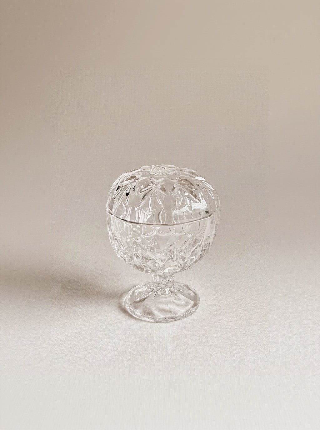 A clear cut-glass or crystal footed bowl with a lid, photographed on a warm beige studio background.