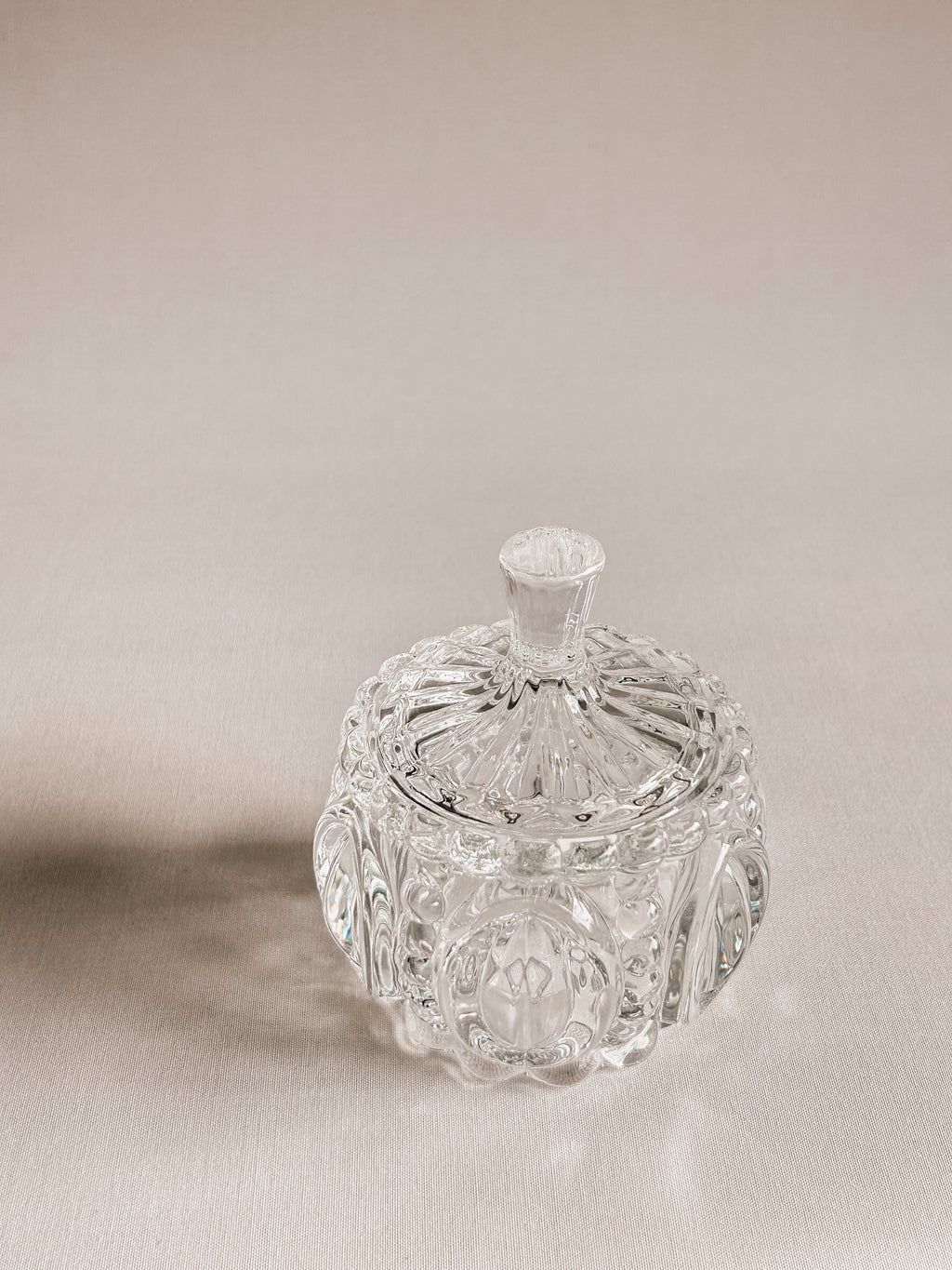 A clear cut-crystal or glass lidded container with ornate faceted detailing and a small top handle, photographed on a warm beige studio background.