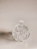 A clear cut-crystal or glass lidded container with ornate faceted detailing and a small top handle, photographed on a warm beige studio background.
