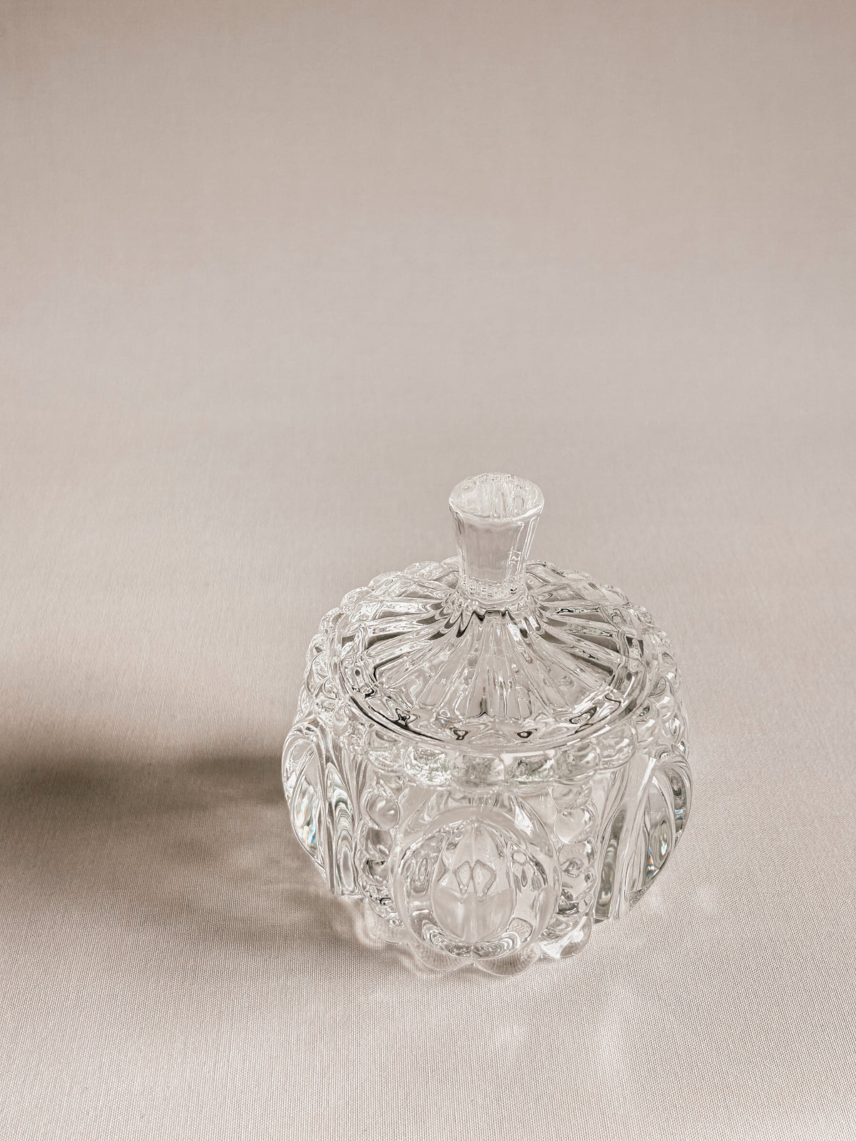 A clear cut-crystal or glass lidded container with ornate faceted detailing and a small top handle, photographed on a warm beige studio background.
