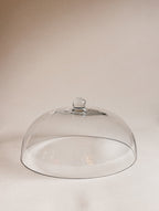 A clear glass cloche dome with a small round knob on top, photographed on a plain warm beige background.