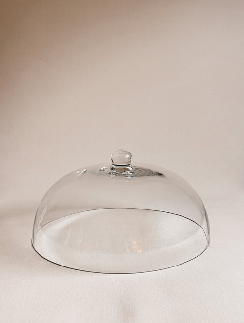 A clear glass cloche dome with a small round knob on top, photographed on a plain warm beige background.