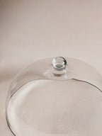 A clear glass cloche dome with a small round glass knob on top, photographed on a soft beige background.