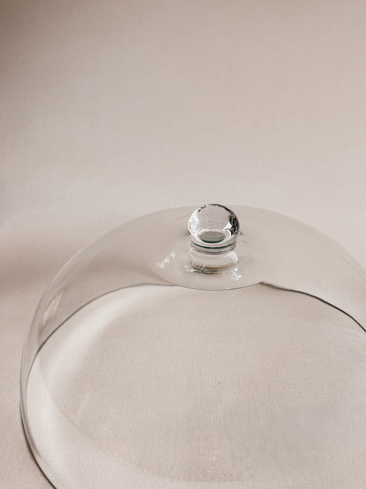 A clear glass cloche dome with a small round glass knob on top, photographed on a soft beige background.