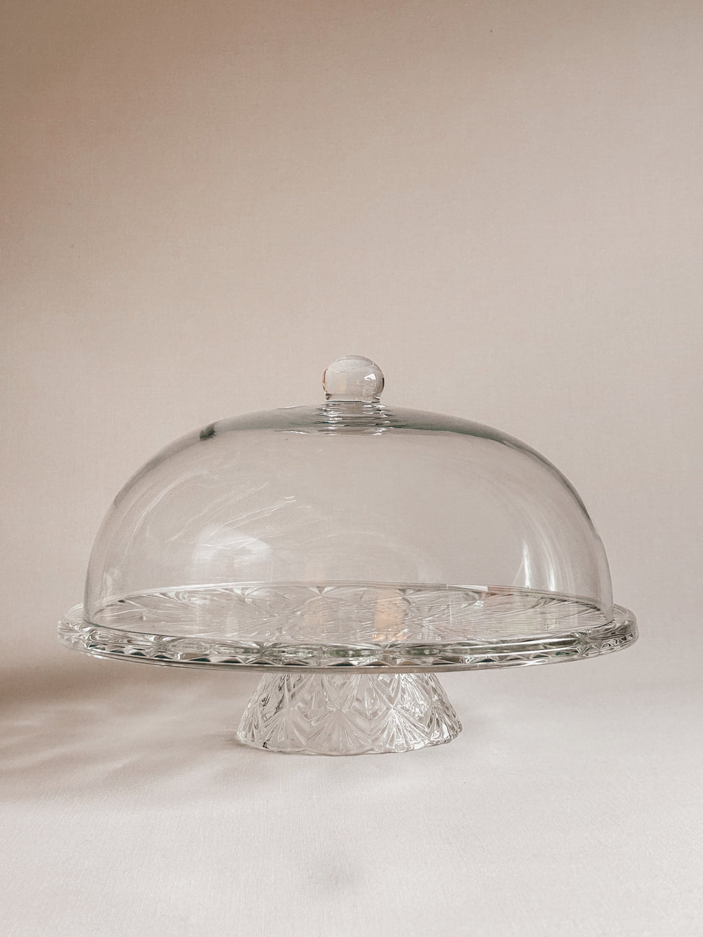 Clear glass cake stand with a matching dome-shaped glass cover and textured pedestal base, photographed against a soft warm beige background.