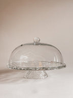 Clear glass cake stand with a matching dome-shaped glass cover and textured pedestal base, photographed against a soft warm beige background.