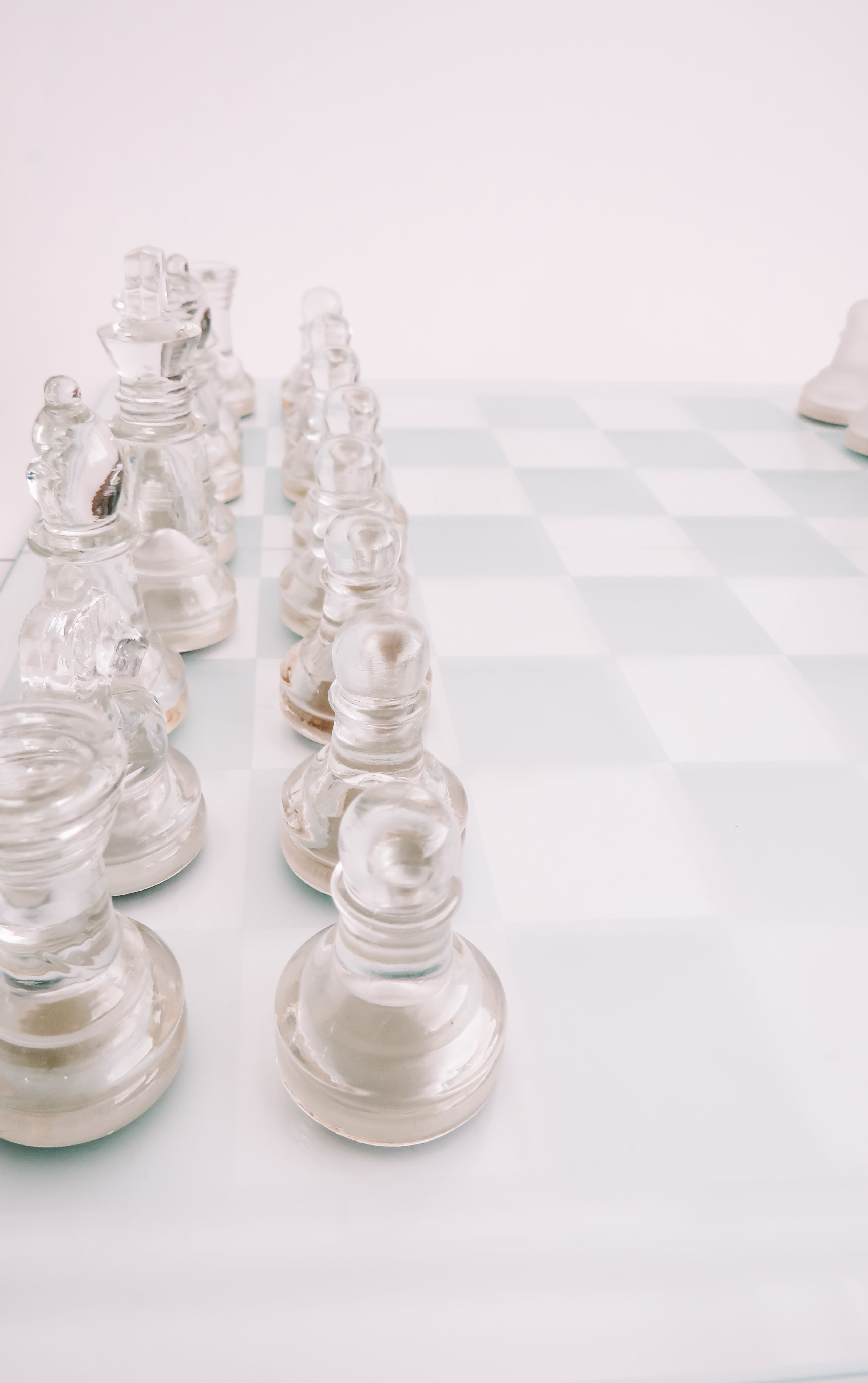 Clear glass chess pieces arranged on a white and pale blue checkered chessboard against a bright white background.