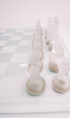 Frosted white chess pieces arranged on a glass chessboard against a clean white background.