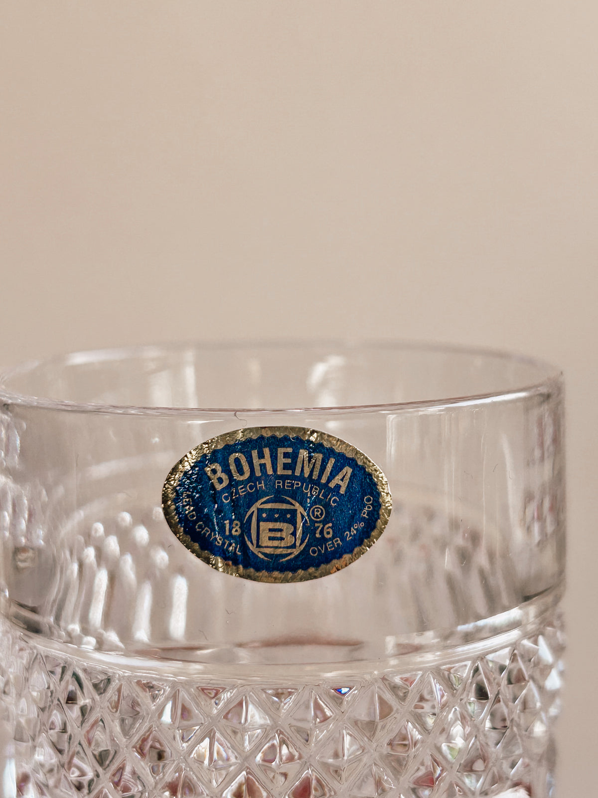 A close-up of a clear cut-glass tumbler with a blue and gold oval 'Bohemia Czech Republic' decal on the side, set against a plain beige background.