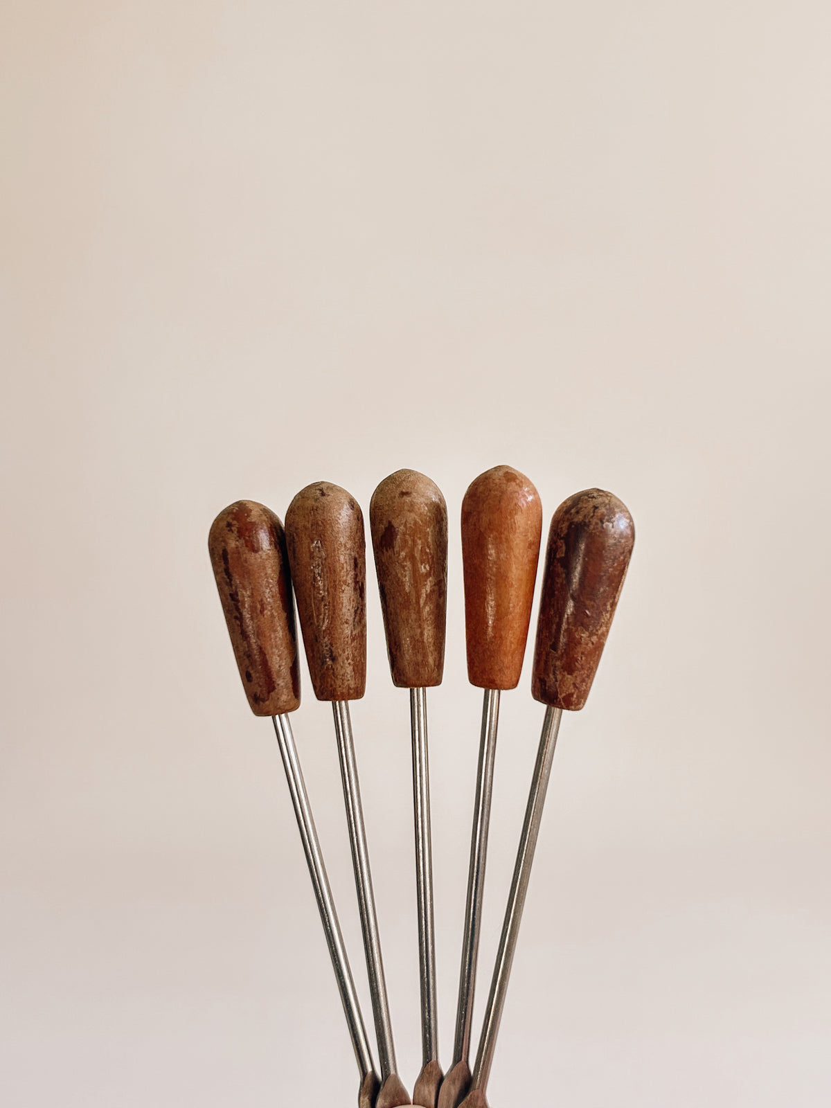 Five wooden-handled metal skewers or tools arranged upright against a plain warm beige background.