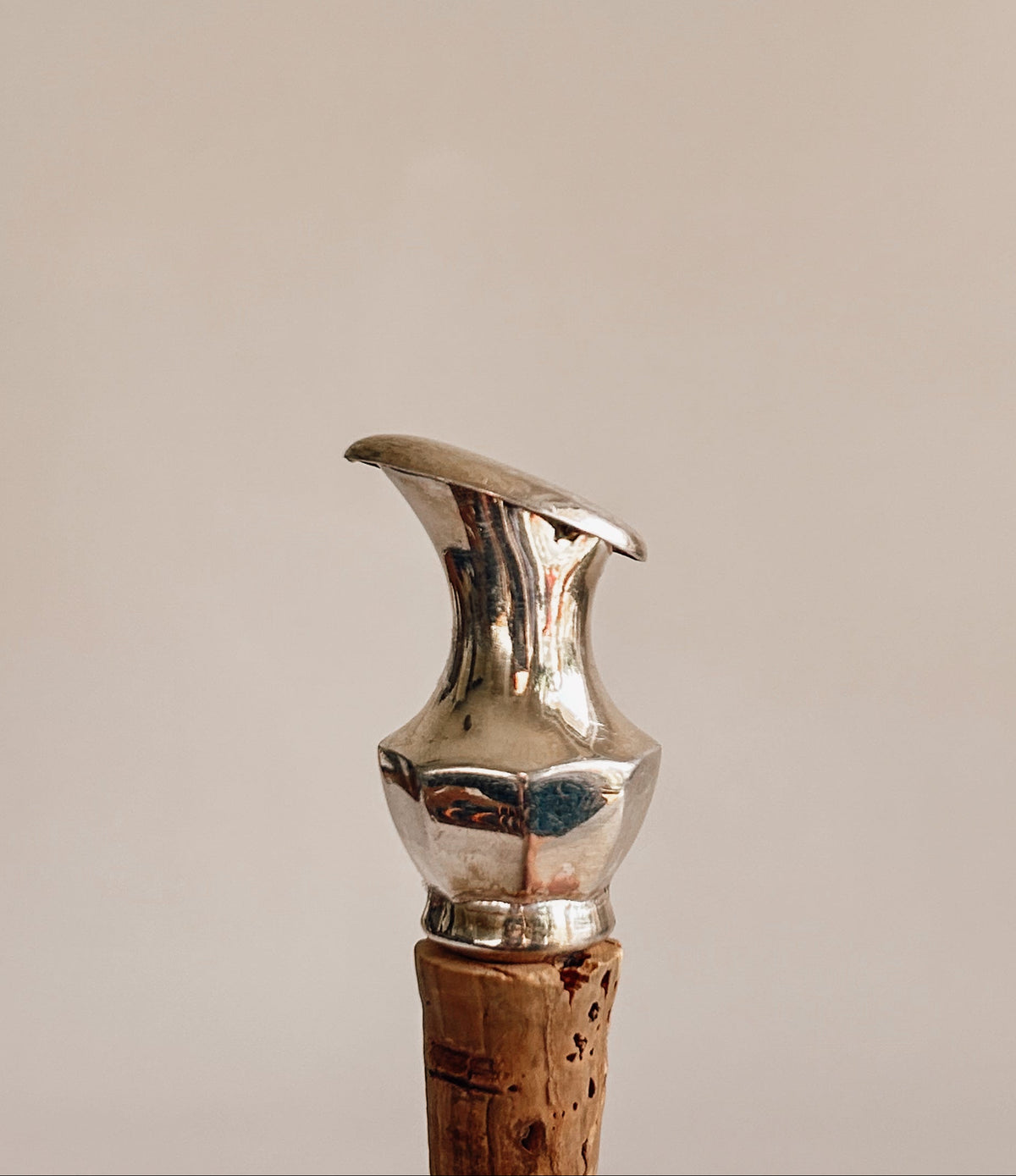 A close-up of a metallic silver corkscrew handle with a cork stopper, set against a solid warm beige background.