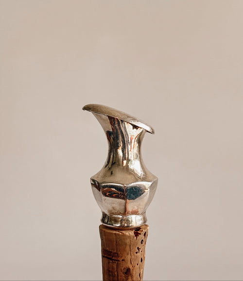 A close-up of a metallic silver corkscrew handle with a cork stopper, set against a solid warm beige background.