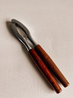 A pair of wooden-handled nut cracker tools with polished silver metal jaws, resting on a light beige textured fabric background.