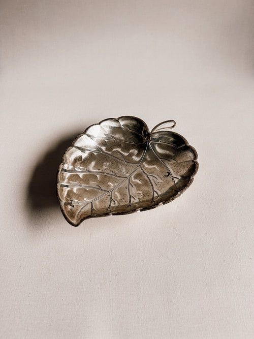 A metallic leaf-shaped tray or dish with engraved leaf veins and a small loop at the top, photographed on a plain light beige surface with a soft shadow.