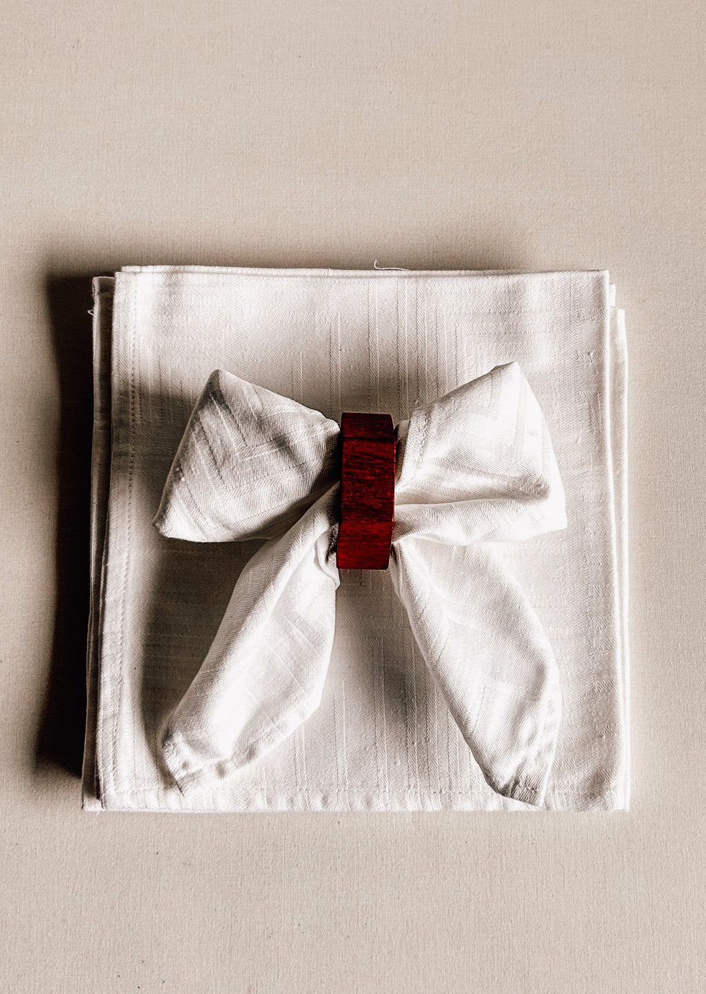 A folded white linen napkin tied in a bow with a dark red fabric band, photographed from above on a warm beige background.